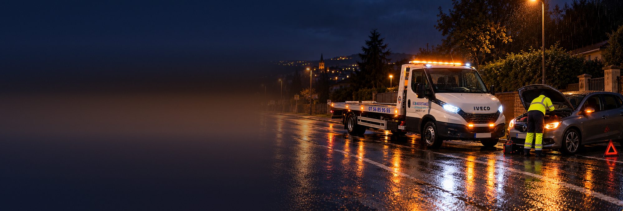 Dépanneur ECO ASSISTANCE démarrant une voiture en panne au booster, de nuit sous la pluie, devant la dépanneuse plateau Iveco Daily, près de Villefranche-sur-Saône